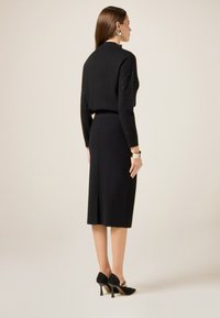 Maglione aderente nero con dettagli in gioiello sulle maniche, abbinato a una gonna nera fino al ginocchio con uno spacco sul retro. Indossato con tacchi neri.