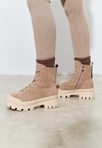 Tan suède enkellaarsjes met dikke beige rubberen zolen, voorzien van veters aan de voorkant en een zijrits, gefotografeerd tegen een neutrale achtergrond.