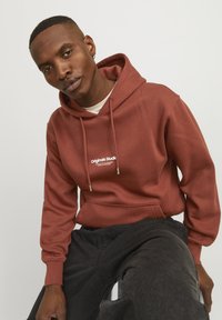 Felpa maroon con cappuccio e lacci, tasca frontale e testo bianco "Originals Studio". Tessuto morbido; abbinata a pantaloni neri.