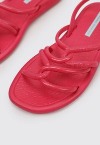 Sandalias de color rosa brillante, hechas de goma, con una plantilla texturizada. Presenta tres correas entrelazadas para un ajuste seguro. Diseño de suela plana.