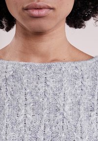 Lähikuva henkilön alalevystä ja kaulasta, joka on pukeutunut vaaleanharmaaseen pitsineulepuseroon, ja kiharat tummat hiukset ovat osittain näkyvissä.