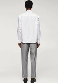 Chemise blanche à manches longues avec un empiècement au dos, associée à un pantalon gris, présentant une coupe slim et des poignets ourlés. Des chaussures noires complètent l'ensemble.