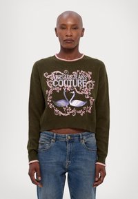 Maglione corto verde con colletto rosa, caratterizzato da cigni bianchi ricamati e scritta "VERSACE JEANS COUTURE", abbinato a jeans blu.