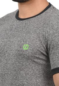 Camiseta gris de manga corta con cuello negro, que presenta un pequeño logotipo bordado en verde en el pecho. La textura parece suave y lisa.