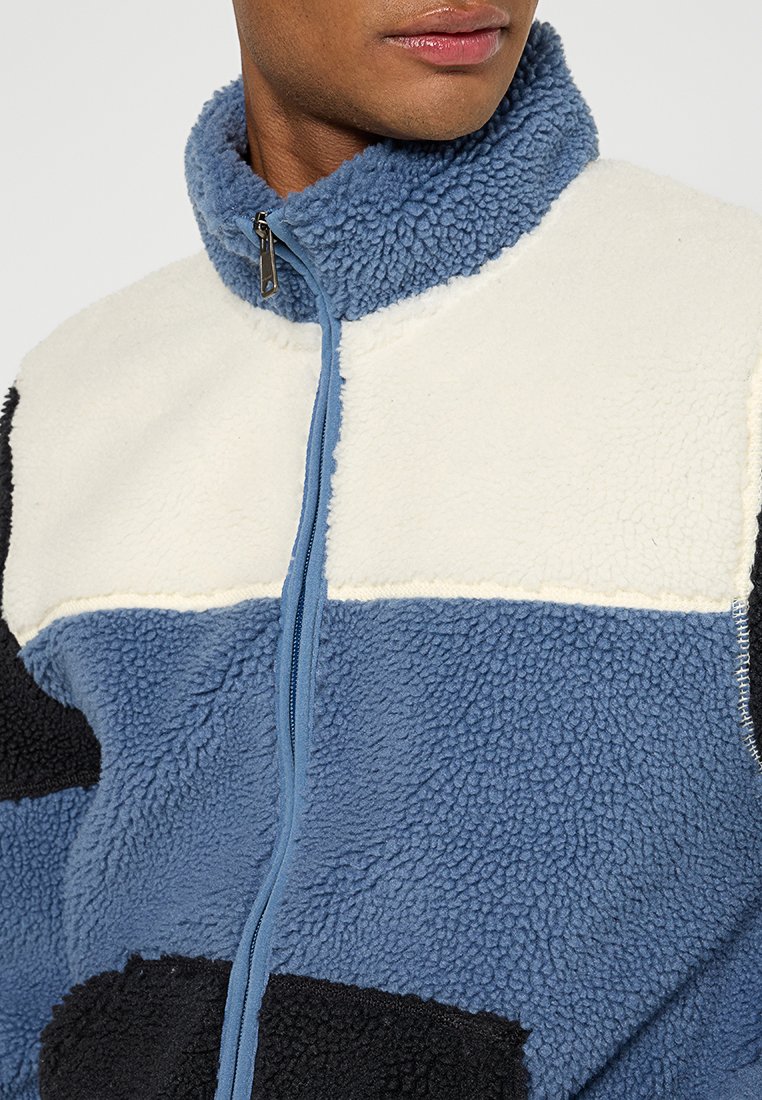 Chaqueta de forro polar en colores azul, blanco roto y negro. Cuello alto, cierre de cremallera y tela texturizada con un diseño de patchwork.