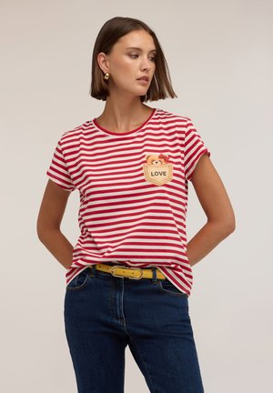Mujer con camiseta de rayas rojas y blancas con un oso de peluche y parche "LOVE", jeans azules y cinturón amarillo, mirando hacia un lado.