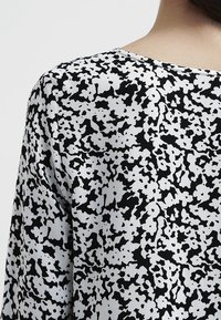 Svart och vit blus med blommönster, mjukt tyg, rund halsringning och avslappnad passform, sedd bakifrån.