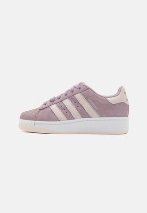 Widok z boku na fioletowo-różowego zamszowego buta Adidas Superstar z kremowymi paskami i grubą białą podeszwą na jednolitym tle.