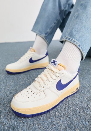 Osoba nosząca białe buty Nike Air Force 1 z niebieskim logo i beżową podeszwą, białe skarpetki i jasnoniebieskie dżinsy na niebieskim dywanie.