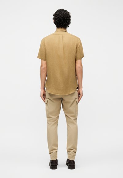 Άνδρας με σγουρά σκούρα μαλλιά, φοράει κοντομάνικο καφέ πουκάμισο, μπεζ παντελόνι cargo και σκούρα παπούτσια, στέκεται γυρισμένος με την πλάτη προς τα εμπρός.