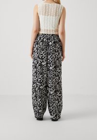 Žena v bezramenném bílém krajkovém topu a volných černobílých kalhotách s abstraktním vzorem, stojící zády k objektivu na jednobarevném pozadí.