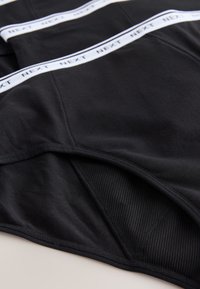 Sous-vêtements en coton noir avec une texture lisse, une ceinture élastique blanche contrastante imprimée à plusieurs reprises du mot "NEXT", et des bords arrondis.