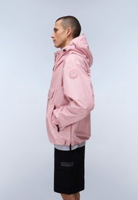 Άνδρας στέκεται προφίλ φορώντας ένα ανοιχτό ροζ αδιάβροχο με κουκούλα, με τα χέρια στις τσέπες και μαύρες βερμούδες, μπροστά σε ένα απλό ανοιχτόχρωμο φόντο.