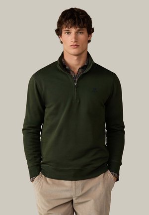 Jonge man die een donkergroene pullover met een kwartrits draagt over een geruit overhemd en beige corduroy broek, staand met handen in de zakken tegen een effen achtergrond.