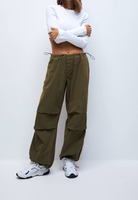 Pantalon cargo kaki en tissu léger et texturé, avec poches latérales, cordons de serrage réglables et coupe ample. Associé à des baskets blanches.