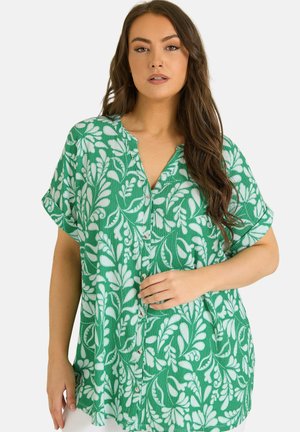 Vrouw met lang bruin haar die een groen overhemd met korte mouwen draagt, bedrukt met witte bladeren, poserend met een hand bij de knopen.