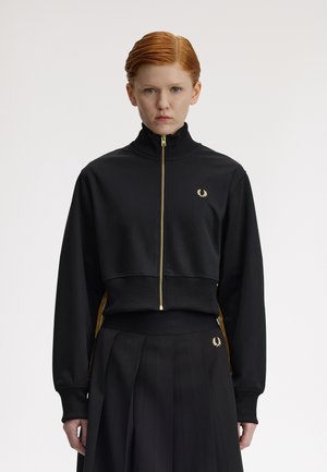 Mujer con cabello rojo que lleva una chaqueta negra con cremallera y una falda plisada, ambas con pequeños logotipos de coronas de laurel doradas en el lado izquierdo.