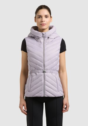 Kobieta w jasnofioletowej pikowanej bezrękawniku z wysokim kołnierzem i paskiem, stojąca na tle jednolitego jasnoszarego tła.