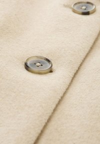 Beigeullt tyg med en mjuk textur, prydd med två stora runda knappar, varav en delvis synlig, med en subtil utskuren design.