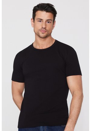 Hombre con cabello corto y oscuro, vistiendo una camiseta negra de manga corta lisa y jeans azules, de pie contra un fondo gris claro.