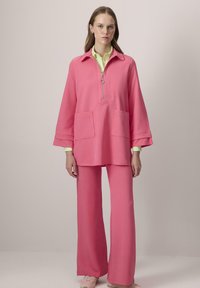 Bluza roz cu fermoar, dotată cu două buzunare frontale, asortată cu pantaloni cu picioare largi. Un tricou galben deschis se vede dedesubt. Textura materialului este moale.