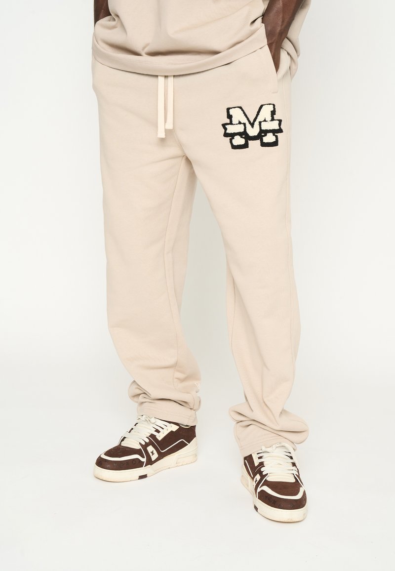 Pantalones de chándal beige con un logo bordado en negro de "M", cintura con cordón y ajuste ceñido, combinados con zapatillas altas en marrón y blanco.