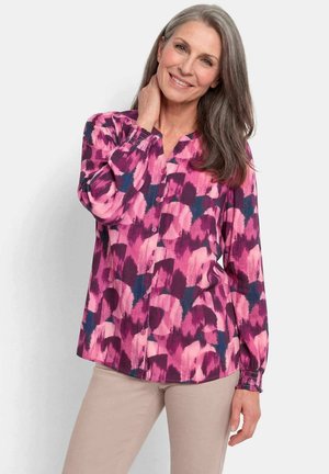 Lächelnde Frau mittleren Alters mit grauen Haaren, die eine rosa und lila gemusterte Bluse mit Knöpfen und hellbeige Hosen trägt, posiert mit der Hand am Hals.