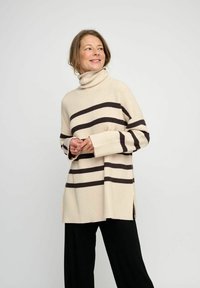 Beige gerippter Rollkragenpullover mit horizontalen dunkelbraunen Streifen, seitlichen Schlitzen und weiten Ärmeln; getragen mit schwarzen Hosen.
