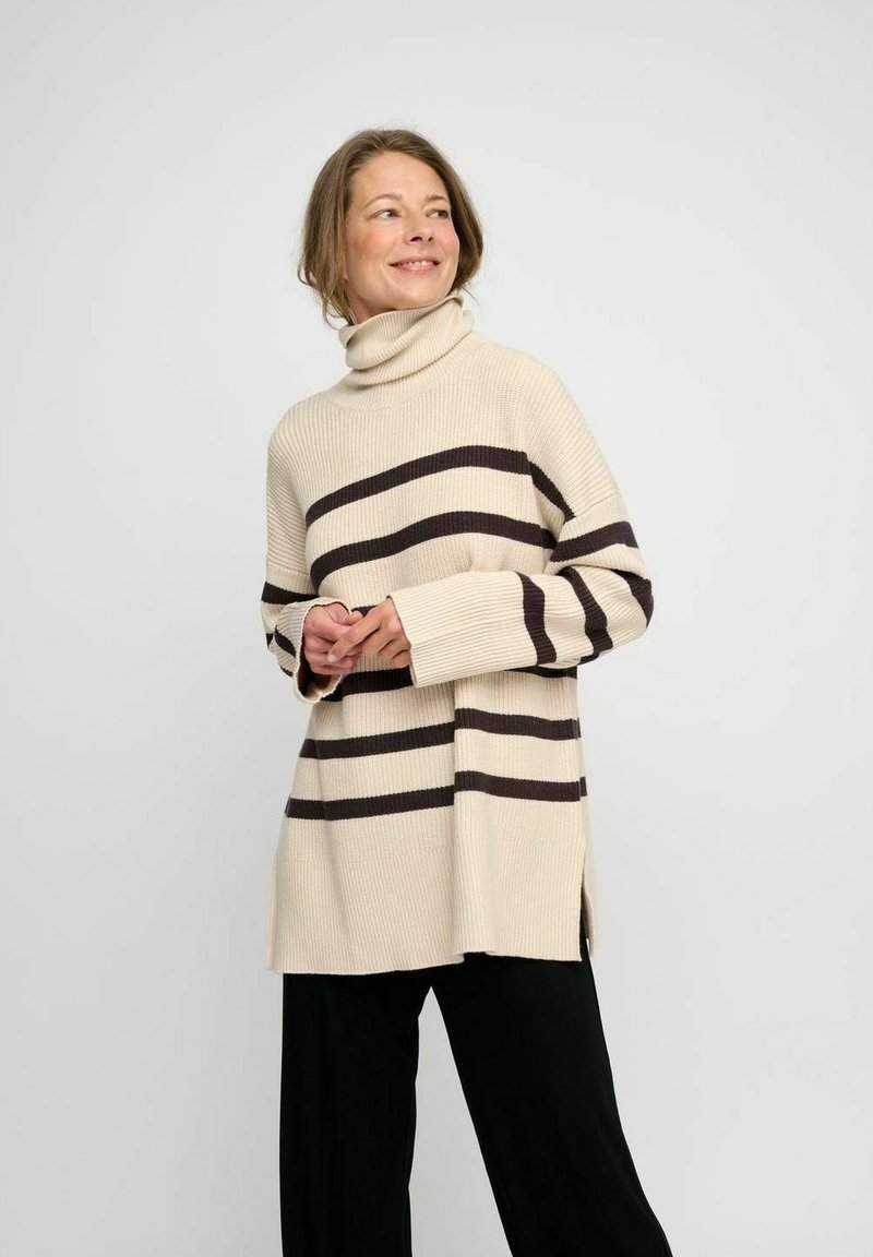Beige gerippter Rollkragenpullover mit horizontalen dunkelbraunen Streifen, seitlichen Schlitzen und weiten Ärmeln; getragen mit schwarzen Hosen.