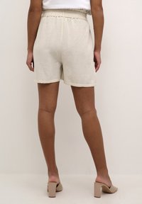 Beige linnen shorts met een gerimpelde tailleband, rechte snit, opgerolde zomen, gecombineerd met beige mules met blokhak op een neutrale achtergrond.