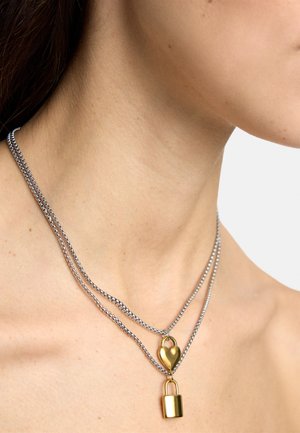 Srebrny podwójny naszyjnik łańcuszkowy z pozłacanym wisiorkiem w kształcie serca oraz złotą zawieszką w kształcie kłódki, noszony na szyi i górnej części klatki piersiowej kobiety.