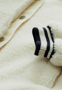 Valkoinen fleece-takki pehmeällä pinnalla, jossa on sileä sisävuori, napit edessä ja ribbireunat, joissa on laivastonsinisiä ja valkoisia raitoja.