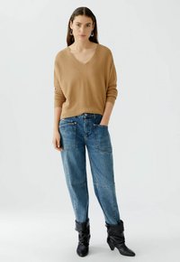 Beige gestrickter Pullover mit V-Ausschnitt, kombiniert mit lockeren blauen Jeans aus Denim mit großen Fronttaschen und schwarzen Knöchelstiefeln.