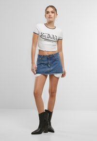 White cropped t-shirt with black "Von Dutch Originals" logo, paired with a blue denim skirt and black heeled boots. Plain background.