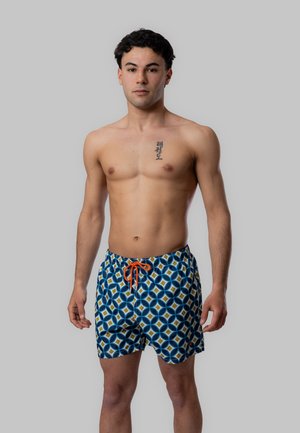 Giovane uomo in piedi a torso nudo che indossa pantaloncini da bagno blu con fantasia e coulisse arancione, espressione neutra, sfondo grigio.