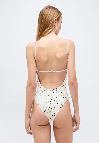 Γυναίκα με μακριά ξανθά μαλλιά που φοράει λευκό ολόσωμο μαγιό με μαύρες πουά, δείχνοντας την χαμηλή πλάτη και τα λεπτά τιράντες του μαγιό.