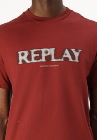 Camiseta de algodón roja con un gran logotipo blanco difuso de "REPLAY" y el texto "NINETEEN EIGHTYONE" en una fuente más pequeña debajo. Mangas cortas.