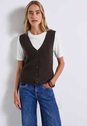 Bruine geribbelde gebreide vest met V-hals en sluiting met vijf knopen, gelaagd over een wit T-shirt. Gecombineerd met blauwe wijde jeans.