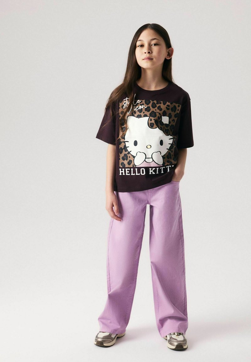 Fille aux longs cheveux debout portant un t-shirt noir Hello Kitty à motif léopard et un pantalon large violet clair sur un fond uni.