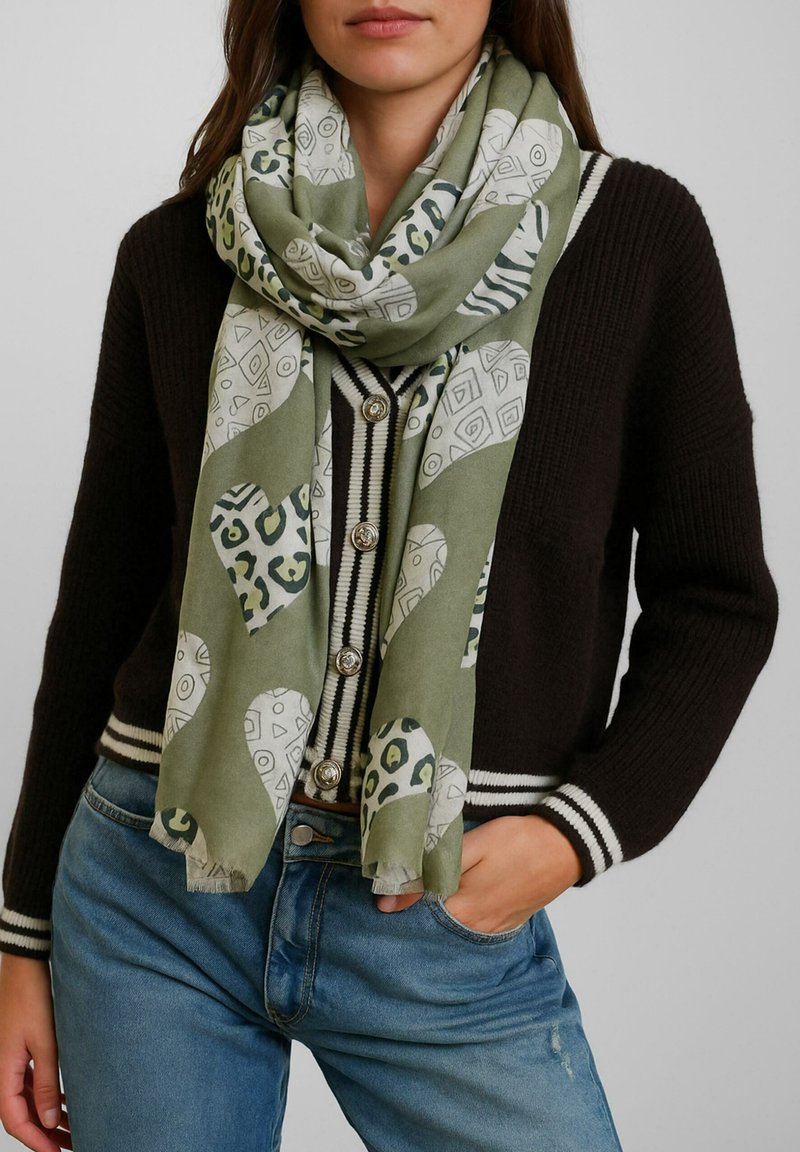 Donna che indossa una sciarpa verde con motivo sopra un cardigan nero con bottoni e strisce bianche, con jeans blu, mano in tasca.
