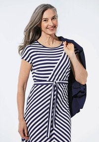 Gestreiftes Kleid in Marineblau und Weiß, mit kurzen Ärmeln, einer taillierten Taille mit Bindegürtel und einem schwingenden Rock. Eine marineblaue Jacke liegt lässig über einer Schulter.