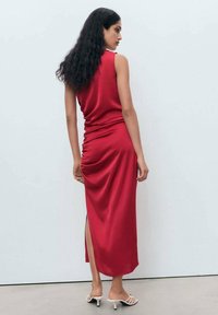 Red satin midi dress with a side slit, form-fitting design, and gathered detailing at the waist. Worn with white strappy heels.