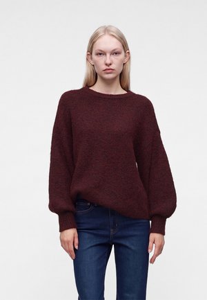 Jeune femme aux cheveux blonds et raides, portant un pull lâche rouge foncé et un jean bleu foncé, se tenant devant un fond blanc uni.