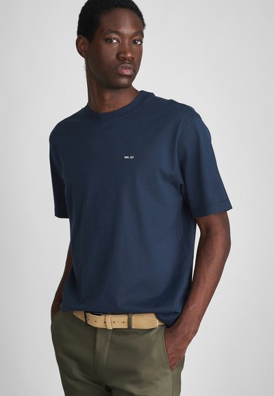 Uomo che indossa una maglietta a maniche corte blu navy con il testo "NN. 07", pantaloni verde oliva e una cintura beige, in piedi con le mani nelle tasche su uno sfondo grigio.
