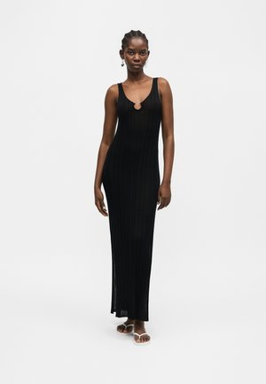 Kvinde iført en sort ribbet maxikjole uden ærmer med en nøglehul-halsudskæring, stående barfodet mod en ensartet hvid baggrund.