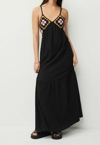 Robe maxi noire à fines bretelles, décolleté en V, et corsage brodé coloré présentant des motifs floraux et géométriques. Texture de jupe fluide.