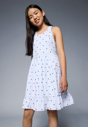 Robe sans manches blanche à rayures bleues et petits motifs de cœurs noirs. Dotée d'un ourlet évasé et d'un corsage ajusté. Texture de tissu douce.