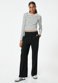 Zwarte wijde broek in combinatie met een aansluitend, langärmig wit shirt met horizontale zwarte strepen. Model draagt grijze sneakers.