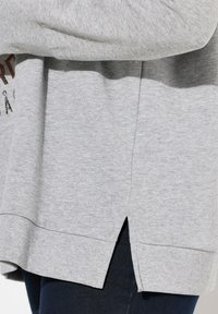 Sudadera gris con un corte relajado, aberturas laterales y puños acanalados. La tela tiene una textura suave y un sutil patrón jaspeado.