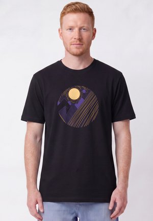 Rothaariger Mann trägt ein schwarzes T-Shirt mit einem runden Grafikmotiv abstrakter Berge, einer gelben Sonne und violetten sowie goldenen Linien.